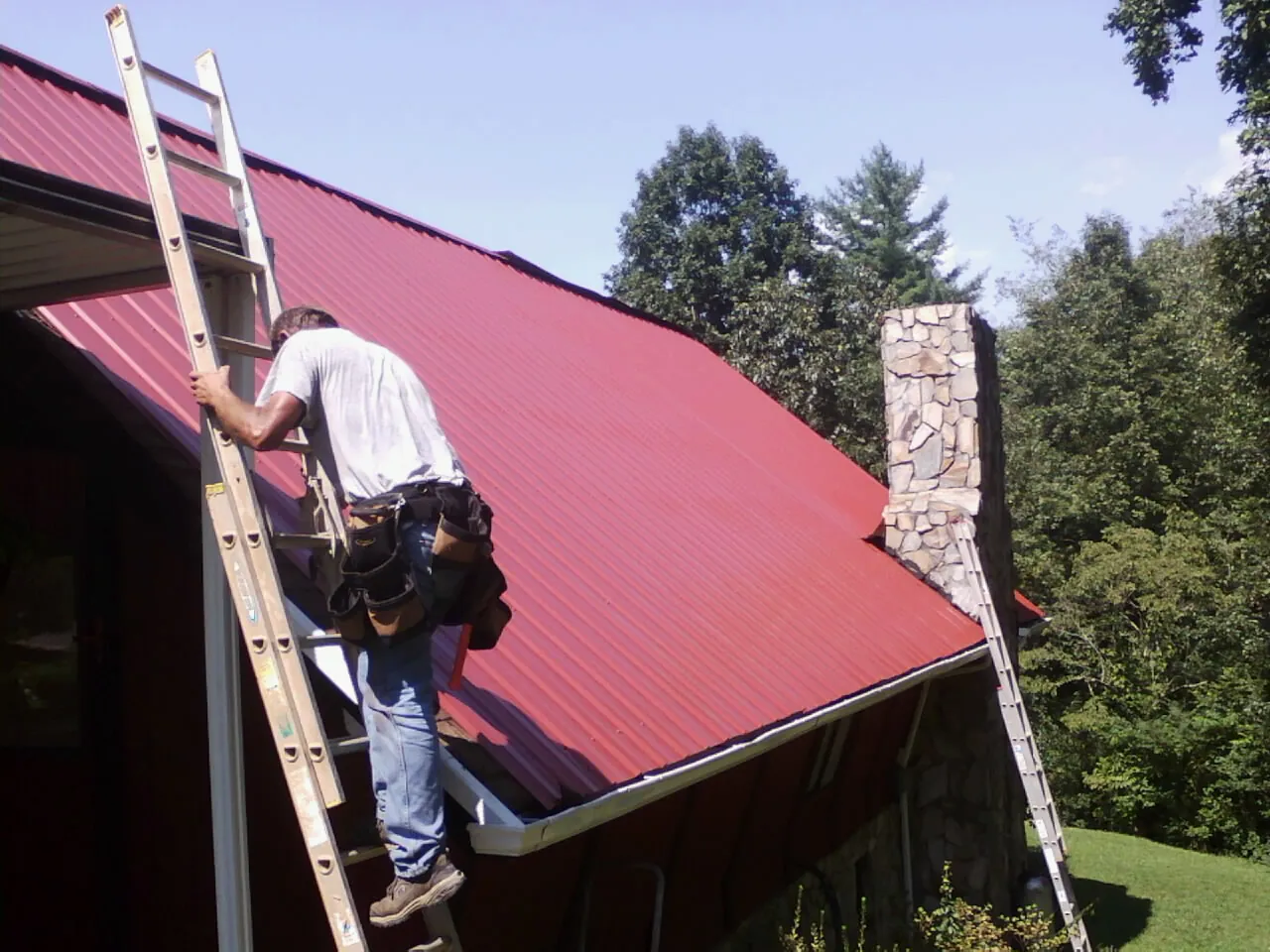 Roofer on a ladder beside a red metal roof and stone chimney — Soffit & Fascia Repair by Roofing Brothers in Washingtonville
